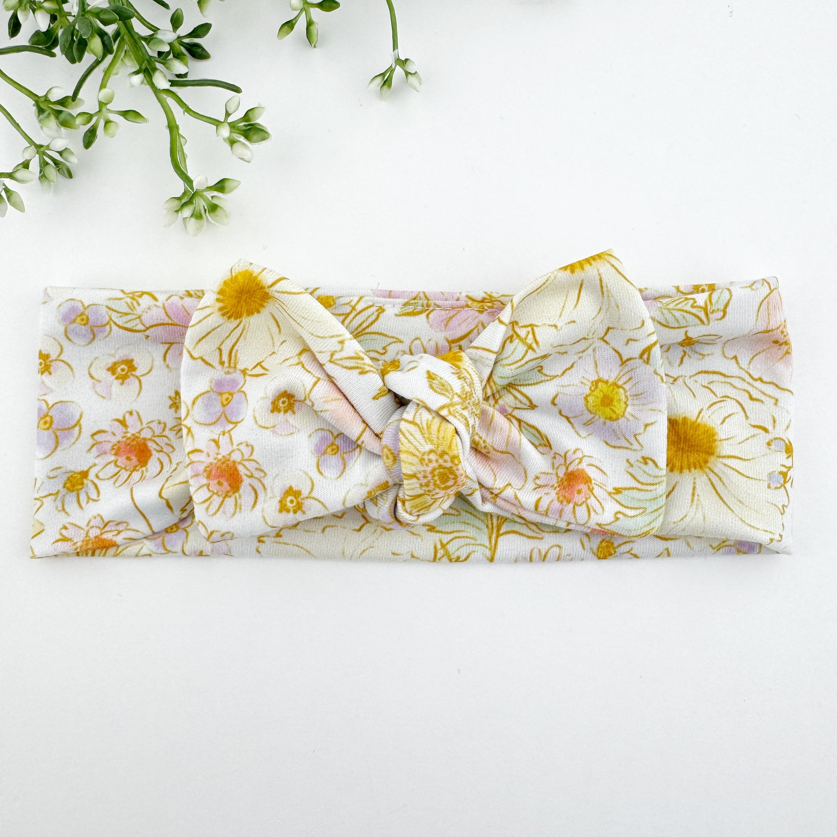 A knotted headband with a floral pattern, featuring a knot in the center, laid flat on a white surface with a plant in the background.