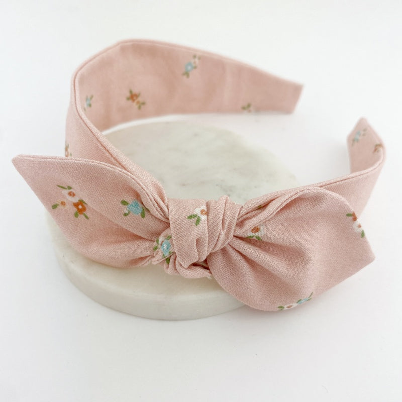 A pink headband with a floral pattern and a removable knot bow, displayed on a white stand.