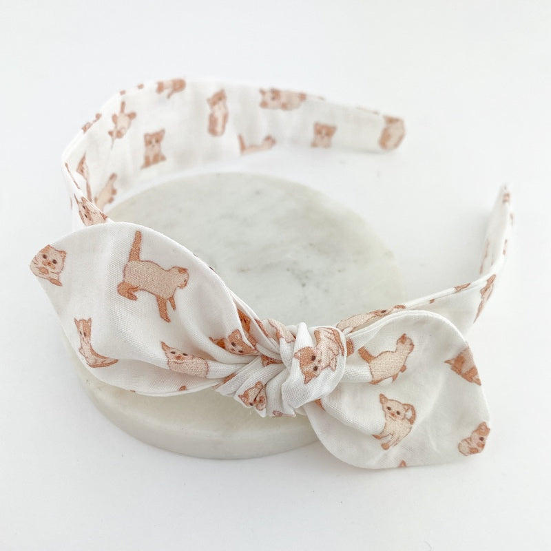 A white headband with a knot bow featuring a pattern of kittens.