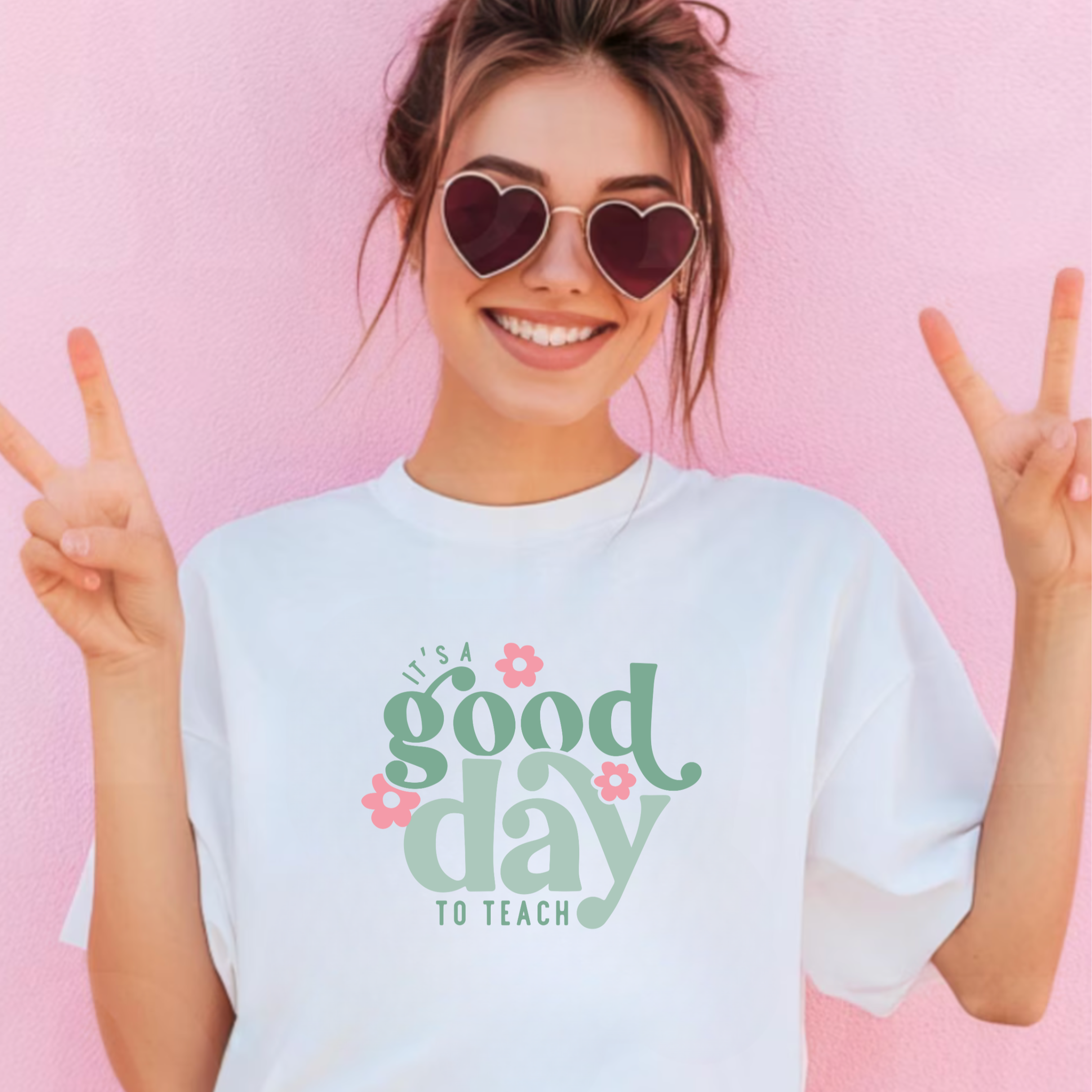 Woman wearing a white t-shirt with 'It's a good day to teach' text and heart-shaped sunglasses against a pink background
