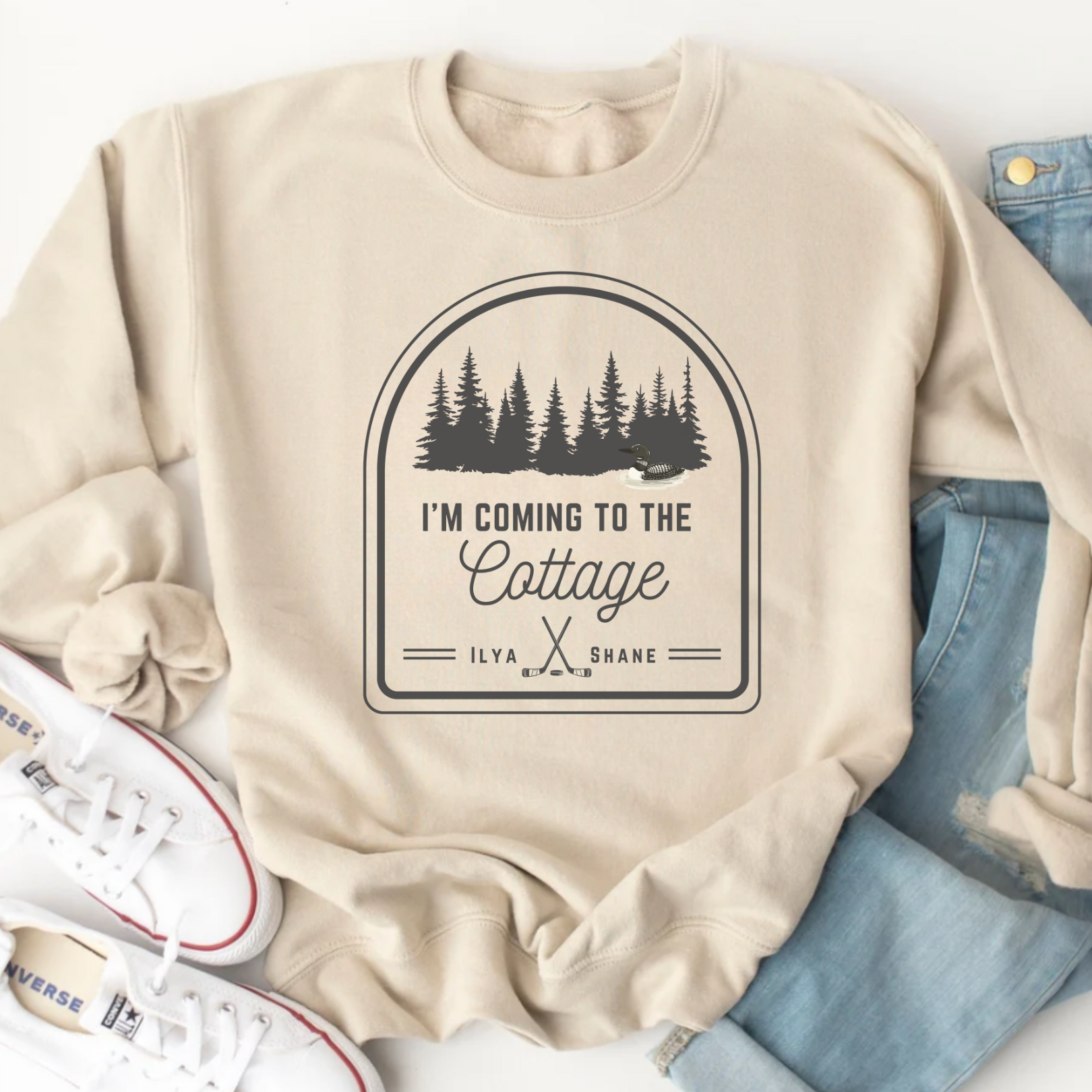 Beige sweatshirt with a graphic design and text, placed on a white surface with jeans and sneakers.