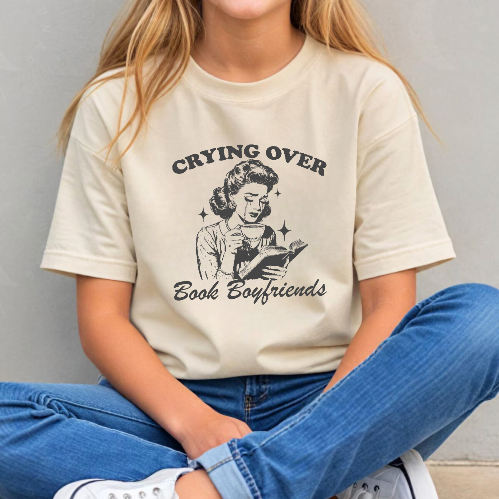 Person wearing a beige t-shirt with a graphic and text "Crying over book boyfriends" 