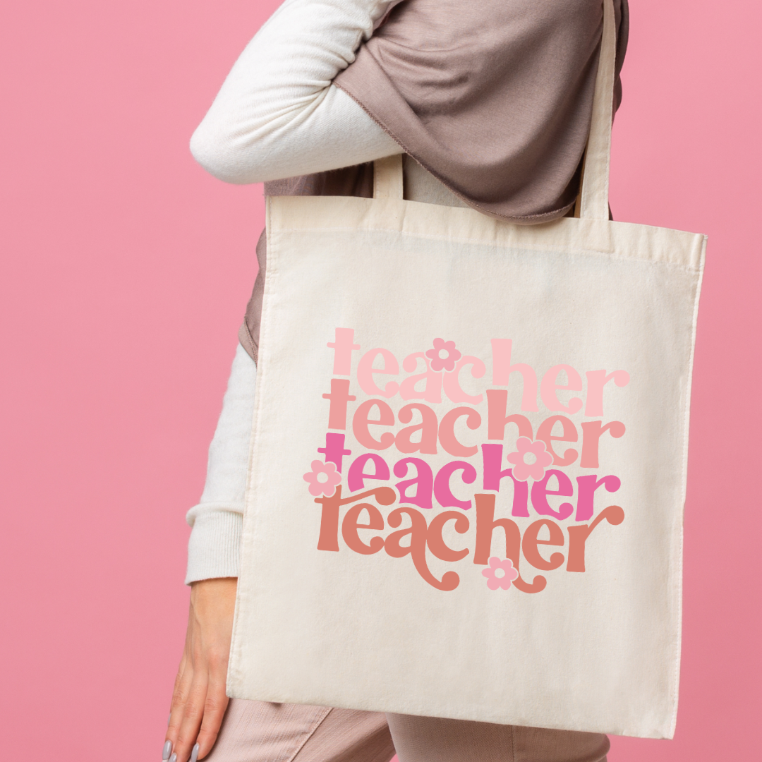 Person holding a tote bag with 'teacher' text in various fonts against a pink background