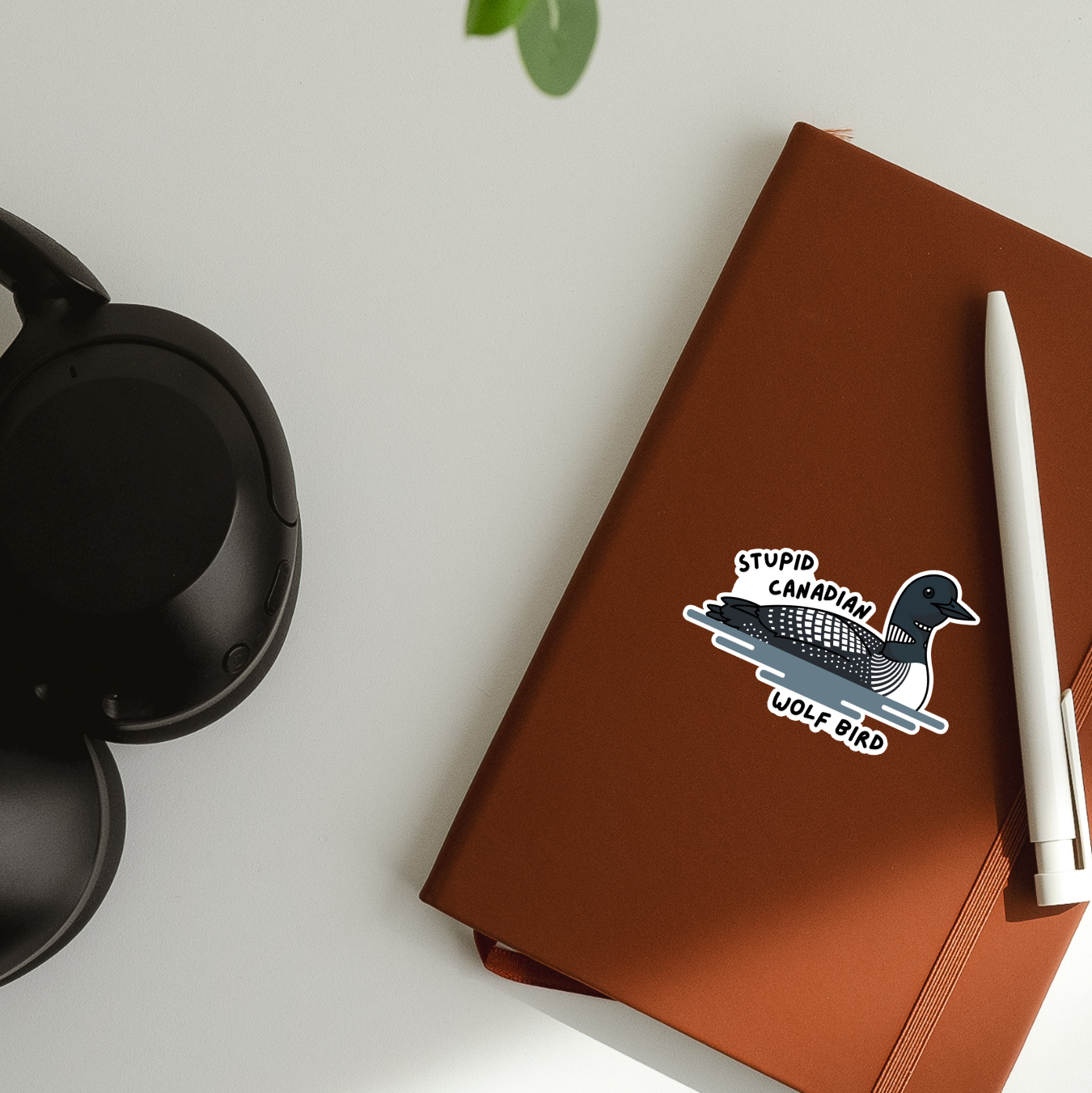 Brown notebook with a sticker on a white surface with a plant and black headphones.